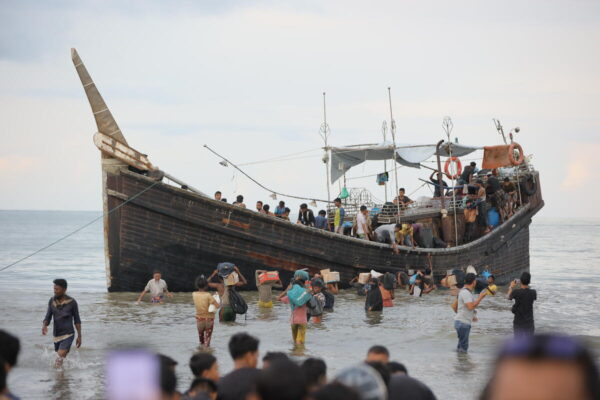 Several people carrying bags on their shoulder and heads, waist deep in sea water. There is a boat in the middle of the image.