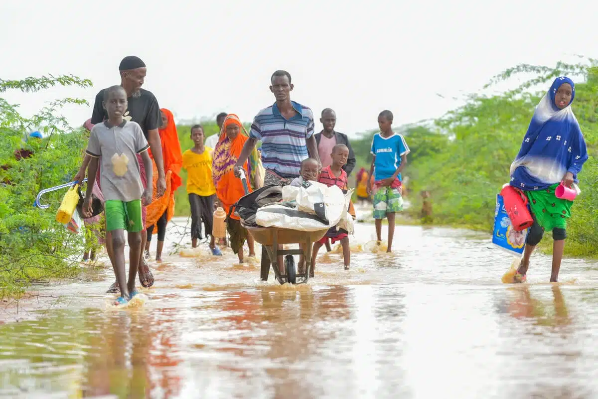 A group of people walking on a flooded street. There is a man in the center of the image pushing a wheel barrel with a child in it.