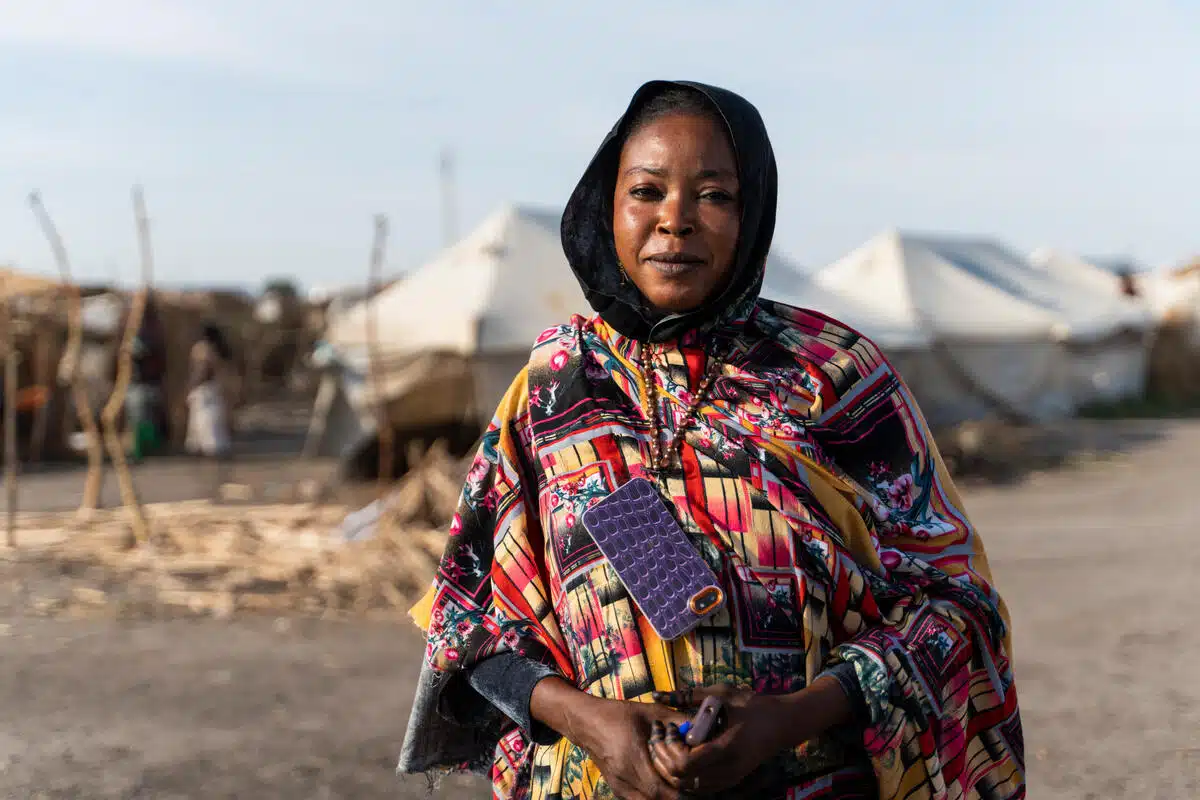 Woman in black headscarf and colourful top is standing in front of three white tents in the background.