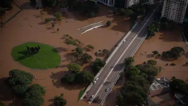 An aerial view of a city experiencing severe flooding. The image shows a bridge over floodwaters, with a few vehicles on it.