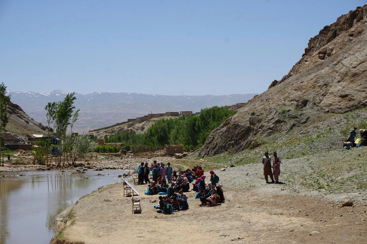 A group of people reunited in an empty area between water and mountains. Some of them are sitting down facing a board. 