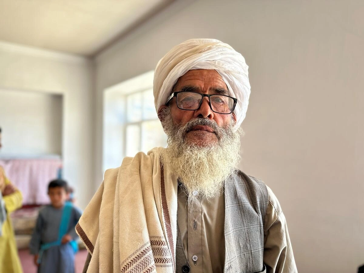 Elderly man standing in front of the camera, looking straight at it. A child is standing behind him.