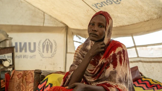There is a woman sitting on a bed in a UNHCR-labeled tent. She is wearing a headscarf.