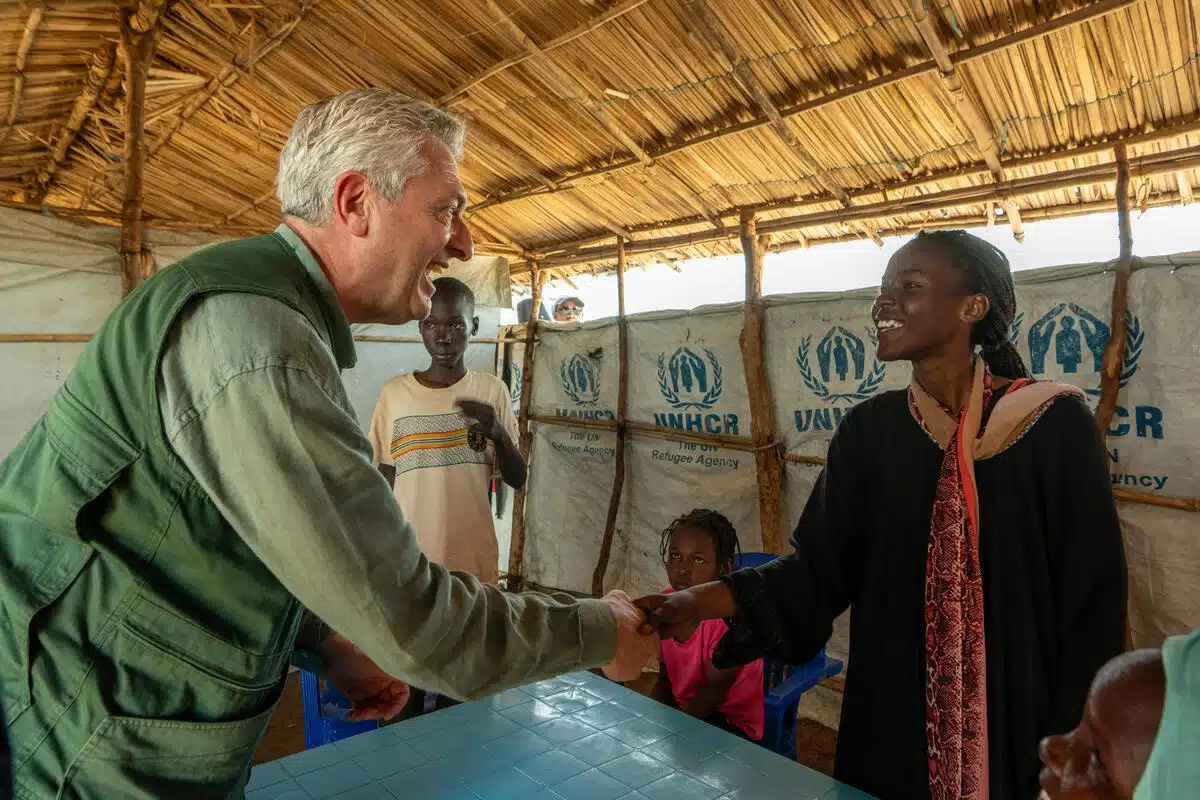 High Commissioner Philippo Grandi is shaking hands with a kid inside a UNHCR-labelled tent. There are other people sitting around them.