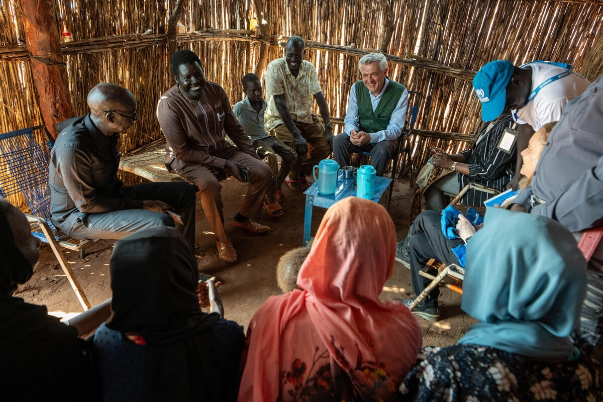 A group of people are sitting in a shelter made of branches. They are smiling and conversating. 