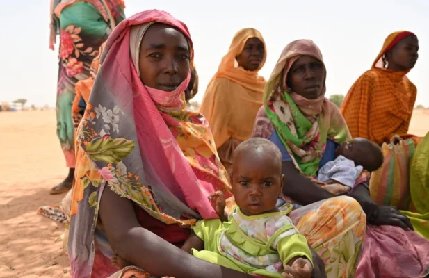 A group of women dressed in colourful clothes and wearing headscarves are sitting on sand. Two of the women are holding babies.