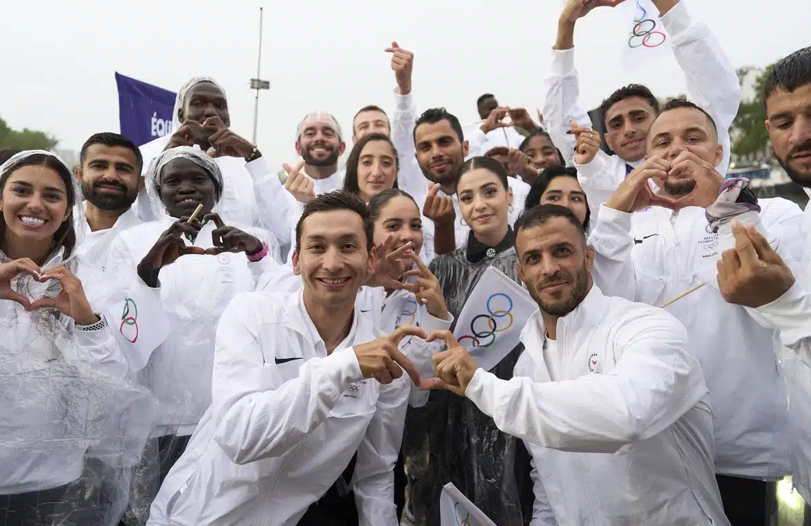 A group of people all wearing white jackets with the Olympics logo are posing for a picture, smiling.