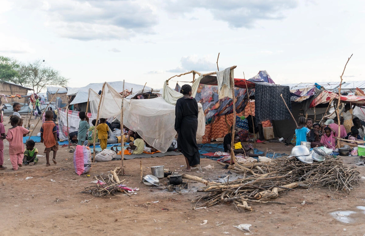 A group of people are in a destroyed area. There are small shelters around them which look incomplete.