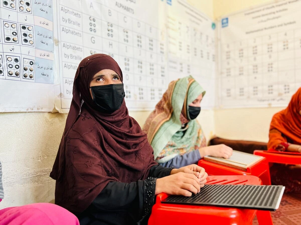 A woman wearing a headscarf is sitting amongst other people with her fingers on a board with small holes in it.