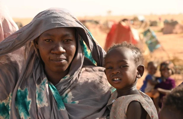 Une femme tient un enfant en bas âge. Ils semblent se trouver dans une zone où il y a beaucoup de sable.