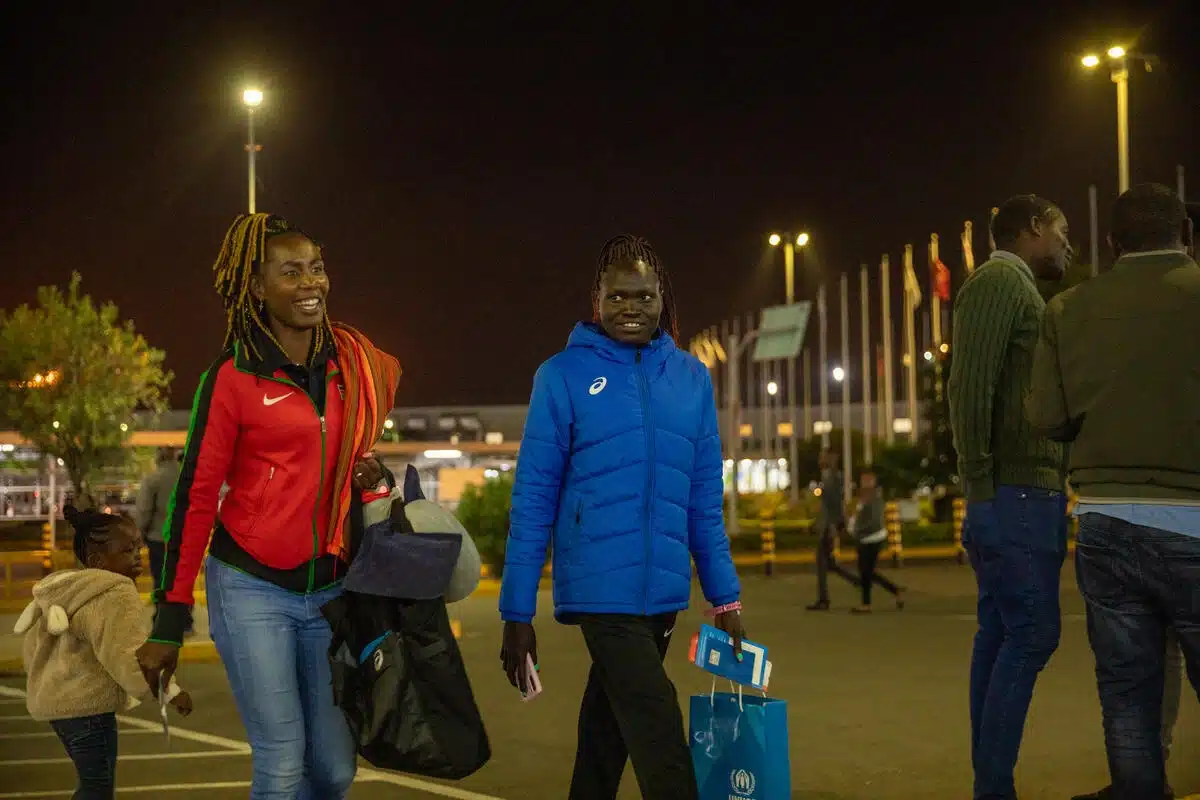 Two women are walking in the parking lot surrounded by tall flags. They are carrying a few bags in their arms. One of them has documents in her hand.