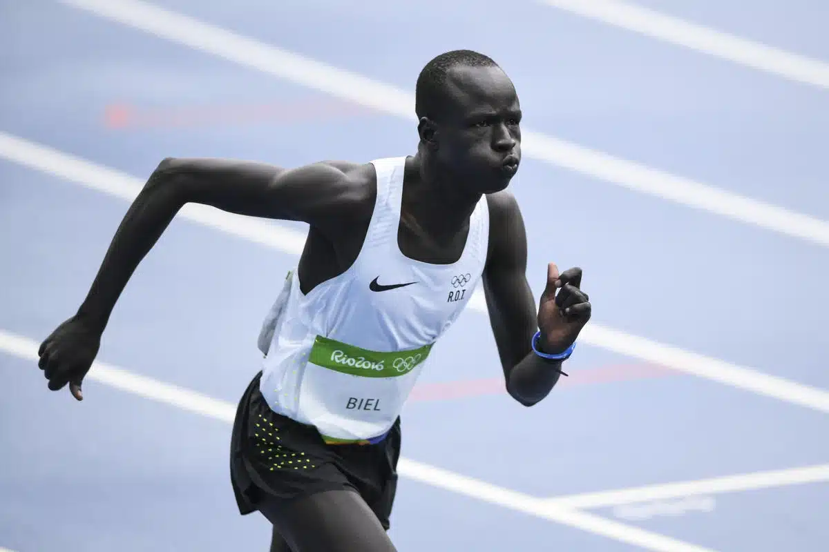 A man is running on a track field. He is dressed in competitive running attire with the Olympics logo on his shirt.