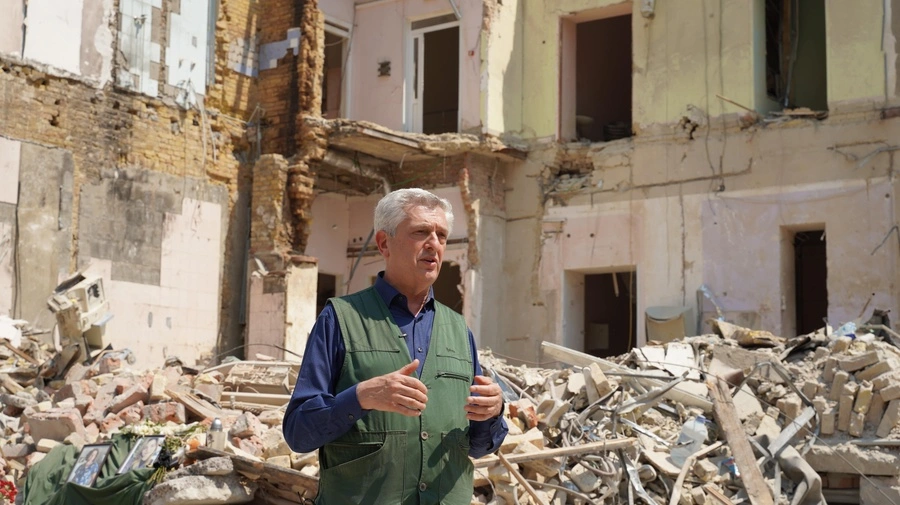 The High Commissioner Filippo Grandi is standing in front of a destroyed building. There is pilled up debris behind him.
