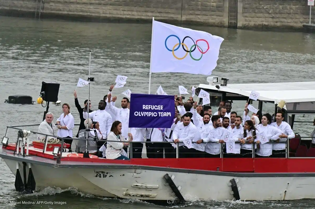 A group of people wearing similar white jackets are on a boat. They are holding a banner that says "Refugee Olympic Team".