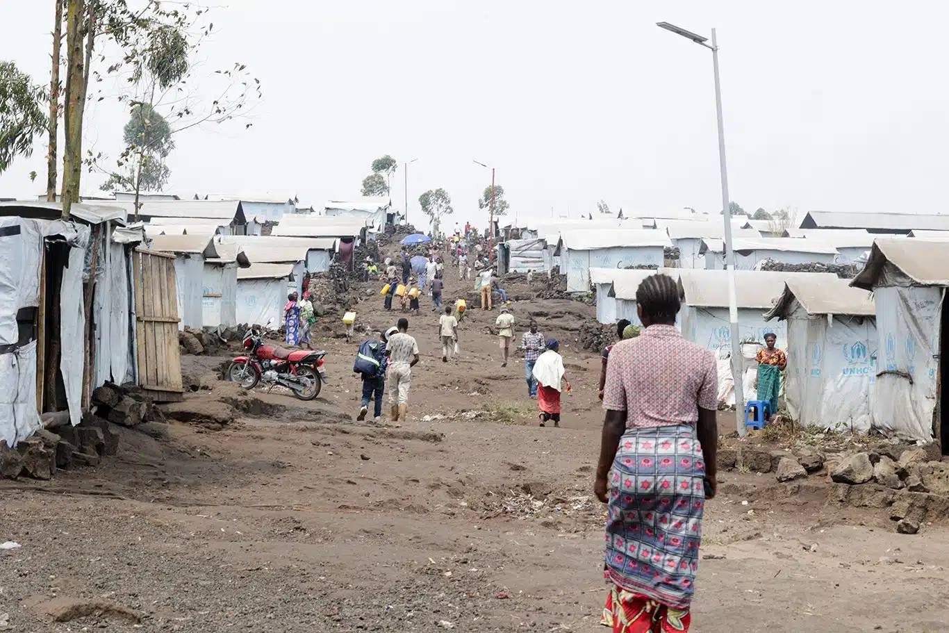 Une femme marche dans une rue boueuse bordée d'habitations temporaires et de tentes recouvertes de bâches en plastique blanc avec le logo du HCR en bleu.