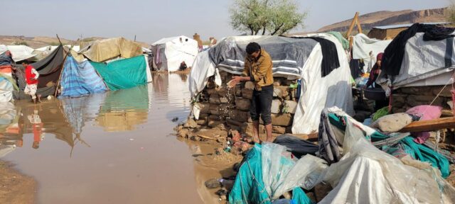 A man in a brown shirt and black pants stands on the side of a large muddy puddle surrounded by flooded makeshift shelters.