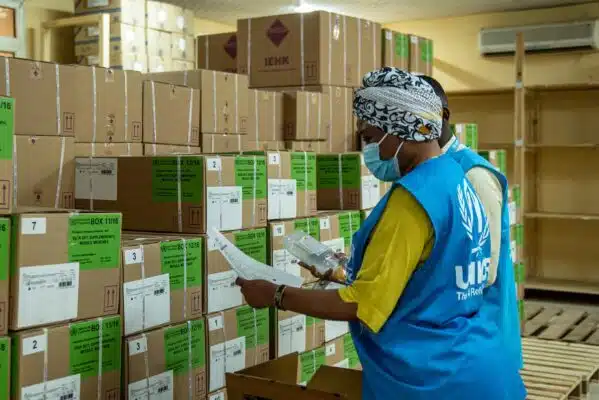 A woman wearing a surgical mask and a blue vest with the UNHCR logo in white examines rows of green boxes at a warehouse.