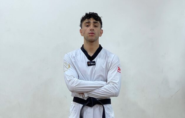 Man in white taekwondo uniform with black collar and belt stands with his arms crossed in front of a white background.