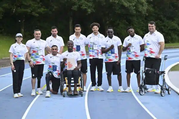 A group of ten people with various disabilities wearing white and blue jerseys and black pants of the Refugee Paralympic Team pose for a photo on a blue and white athletics track.