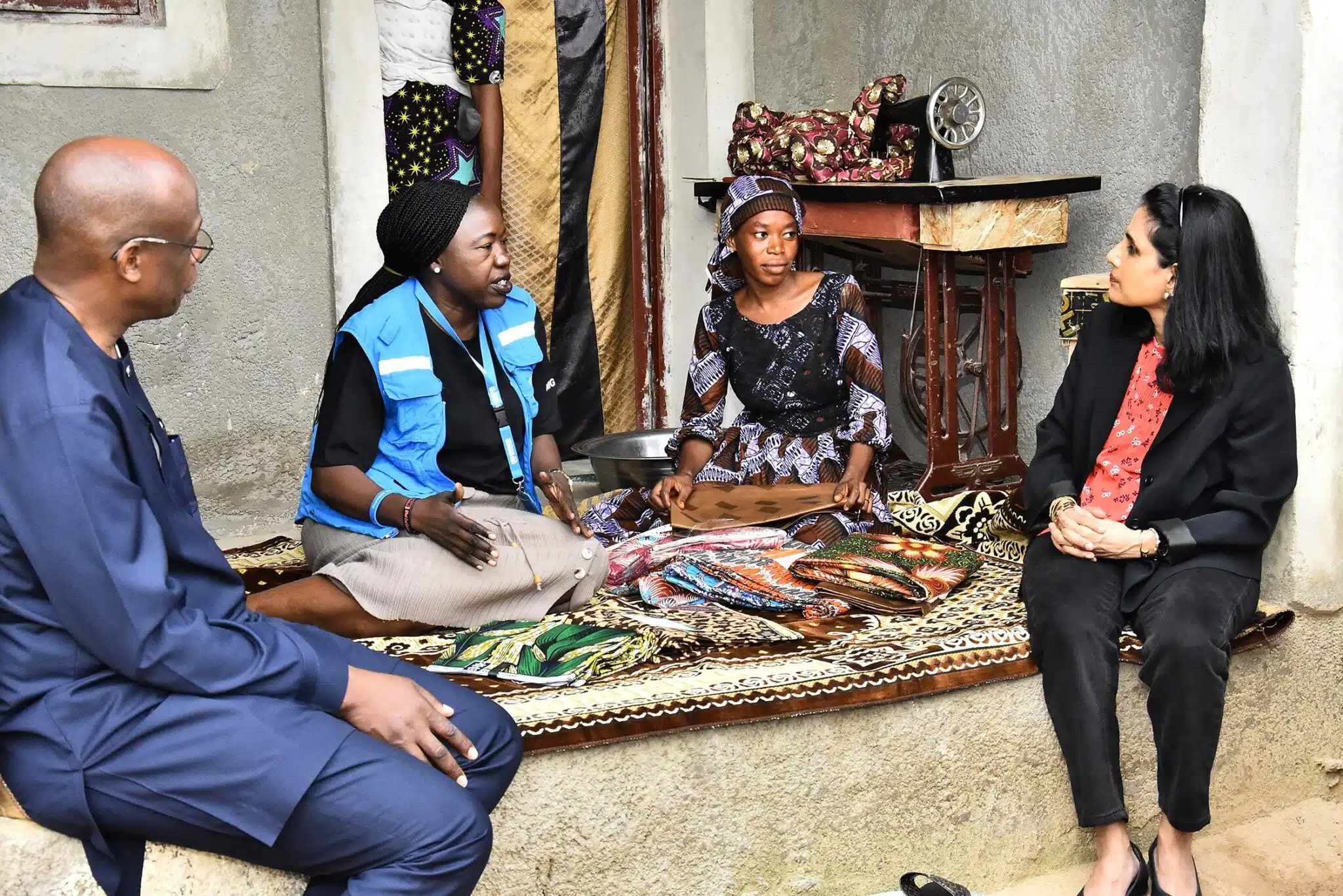 Un homme et trois femmes discutent, assis sur un sol surélevé recouvert d'un tapis et de tissus. Une jeune femme africaine vêtue d'une robe brune et blanche et d'une coiffe assortie est assise à côté d'une vieille machine à coudre à pédale et montre des morceaux de tissus colorés à une femme sud-asiatique vêtue d'un tailleur pantalon noir et à une femme africaine portant un gilet bleu du HCR, tandis qu'un homme noir vêtu d'un costume bleu de style africain est assis sur la gauche.