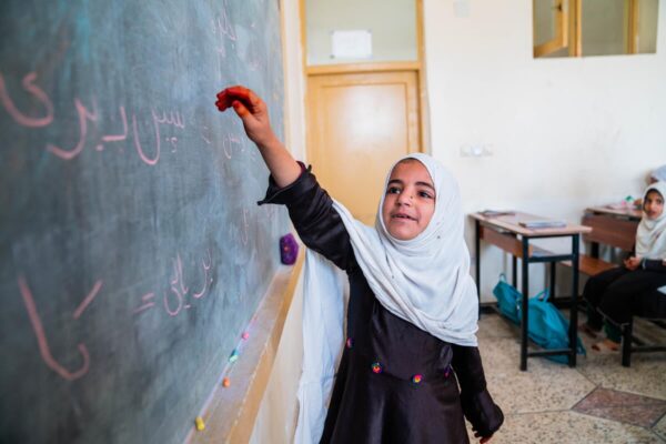 A young girl wearing a white hijab that covers her head and shoulders and a black school uniform dress writes on a classroom blackboard with pink chalk letters in Pashto language.