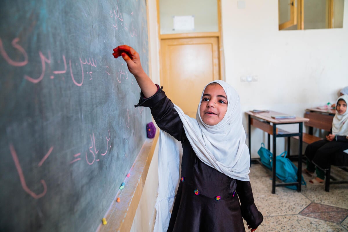 Une jeune fille portant un hijab blanc qui lui couvre la tête et les épaules et une robe d'uniforme scolaire noire montre de la main droite un tableau de classe avec des lettres à la craie rose en langue pachtoune.