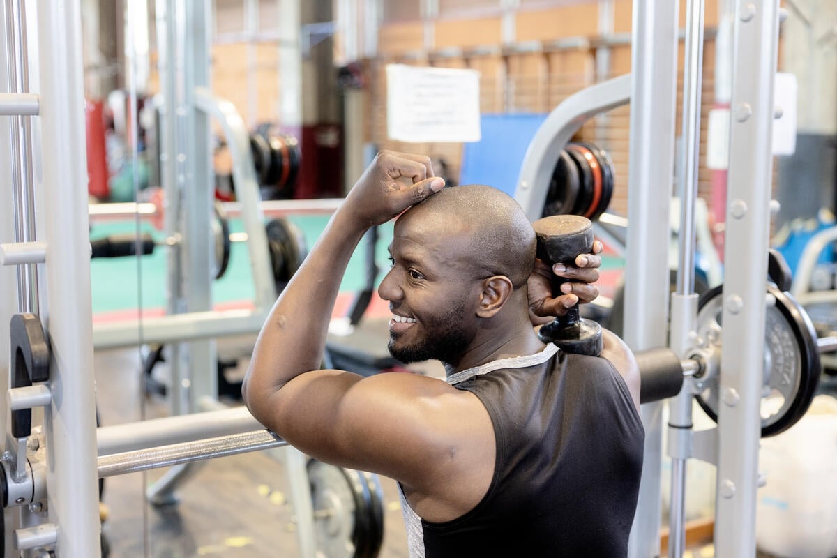 Une personne assise dans une salle de sport sur un banc de musculation, reposant le poids soulevé dans sa main droite et sa main gauche au-dessus de sa tête. Un miroir occupe la majeure partie de l'espace mural à l'avant de la salle.