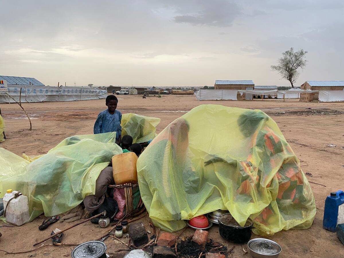 A refugee boy stands in a muddy desert field with bright yellow plastic sheets covering the family's belongings strewn on the ground and the outlines of two women visible under one of the plastic sheets as they try to shelter from rain. Large temporary structures covered with white plastic sheets with light blue UNHCR logos on them can be seen in the background.