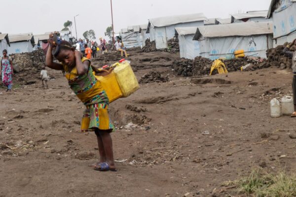 A child wearing vibrantly coloured dress carries two jerrycans with a bent-back and a rope over the top of their head padded with a sandal. In the background are several shelters with the UNHCR logo displayed on the tarping around them. The environment is muddy with no greenery and cloudy skies.