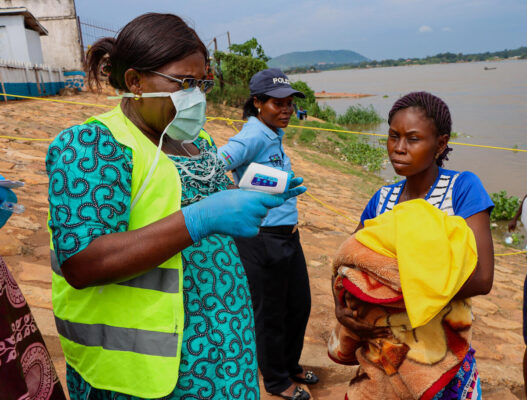 A black woman wearing a medical mask and surgical gloves holds an electronic thermometer in her right hand as she prepares to take the temperature of another black woman holding a bundle of blankets and a bright yellow piece of fabric as they stand on the shore of a body of water with a black female police officer looking on.