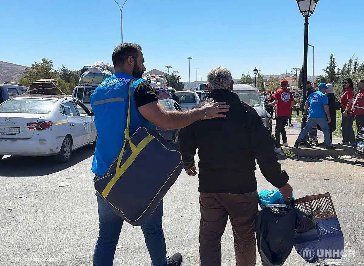 A person wearing a blue UNHCR vest carries a blue and yellow duffle bag while walking with their hand on another person's back, the person is carrying miscellaneous belongings.