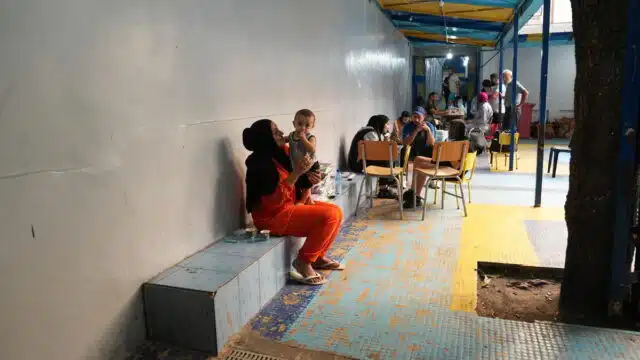 Person wearing a black head covering and orange full body clothings holds a baby in an area covered with colorful ceramic tiles and dirt. People gather in the background with body language that may be taken as concerned.