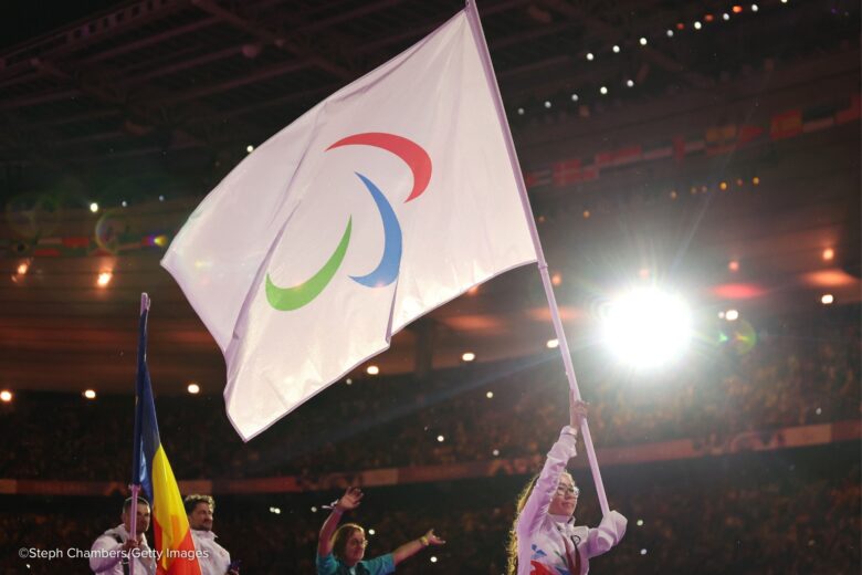 Porte-drapeau brandissant le drapeau du CIP. Un stade rempli de monde remplit l'arrière-plan, tandis qu'une lumière stroboscopique apparaît dans le coin supérieur droit.