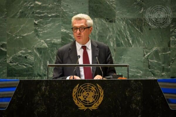 UN High Commissioner for Refugees Filippo Grandi looks toward the camera as he delivers a speech. He stands behind a black marbled podium that displays the UN logo in a gold overlay.