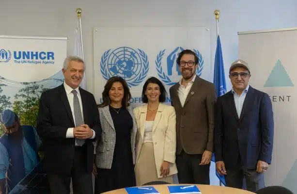 The High Commissioner stands around a table with four other people that are additionally professionally dressed. The room is filled with flags and banners displaying the UNHCR logo.