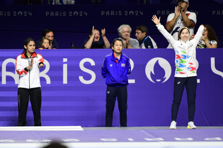 Des athlètes paralympiques se tiennent sur une plate-forme devant une foule en liesse. Les deux athlètes de gauche sont sans expression tandis que le troisième athlète de droite applaudit.