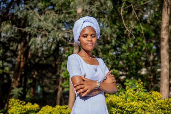 Woman stands in a light blue dress and headwear with her arms crossed. She looks at the camera while the background is filled with trees and greenery. The sunlight shines on her face.