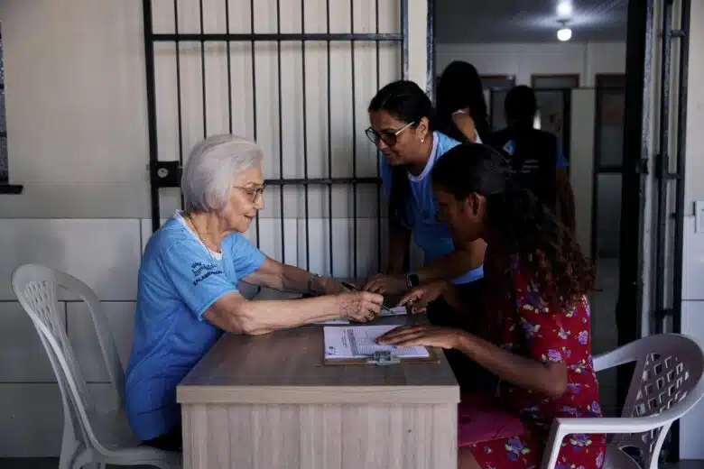 An elder woman points to documents on a desk as two women look towards where she points. 