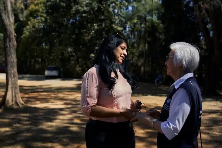 Two women stand in a garden. The older woman on the right embraces to hands of the woman on the left as they stare at each other compassionately. 