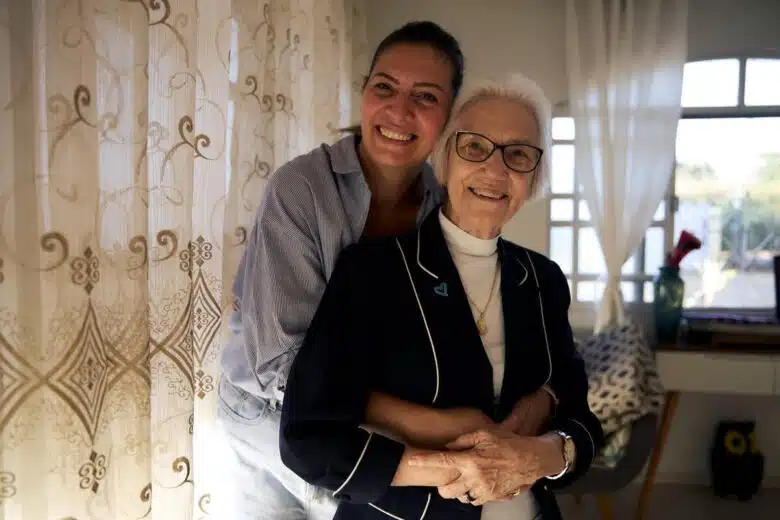 Two women embrace while looking at the camera in a room lit by natural light with light coloured curtains and furniture.