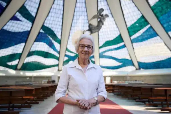 Woman stands holding her hands, interlocked in front of her inside of a building with a stained glass, dome-shaped ceiling. An angel is suspended from the ceiling and pews line the room.