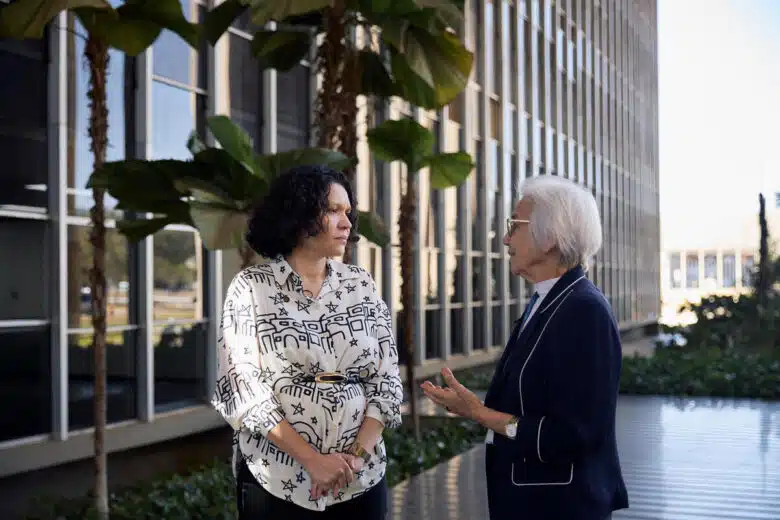 Two women stand in front of a large building with many windows. They are in conversation, the woman on the right is presenting her hands towards the woman on the left. The woman on the left is holding her hands intertwined in front of herself.