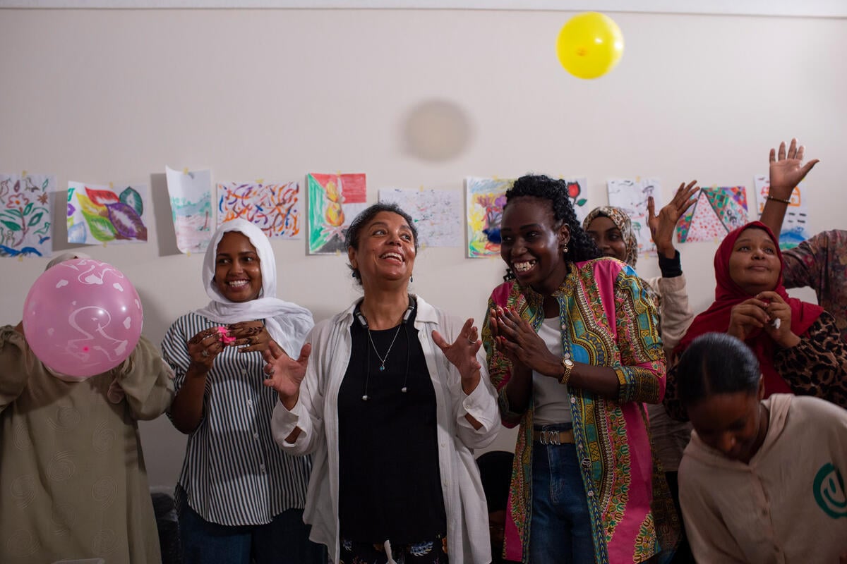 Seven women celebrate with balloons as they cheer in front of a wall lined with papers that display drawings.