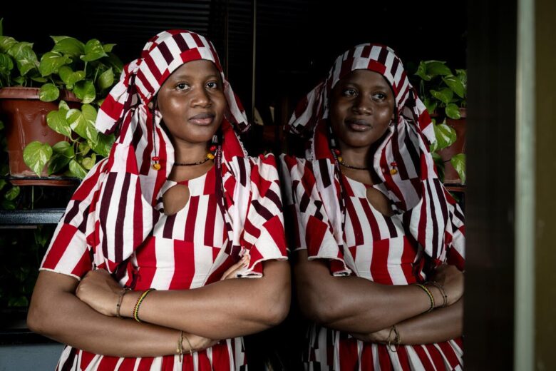 Woman wearing a red and white striped outfit and head covering leans against a mirror while looking into the camera. The room is dark and a plant is in the back.