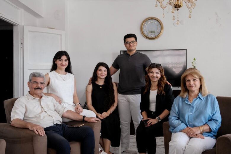 An older man and woman sit at the front of the left and right side of a family room. A woman in white sits on the edge of a sofa next to the older man on the left. Next to the older woman on the right but slightly behind, are two women wearing black. The one on the left is in a dress and the one on the right is wearing a blazer. A man wearing white pants and a dark shirt stands at the back with his arms open.