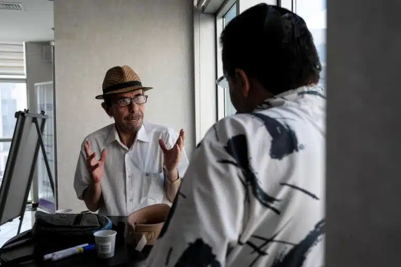 Two men have a conversation. The camer is focused on one who is talking expressively with his hands and the other appears to be listening, his back towards the camera.