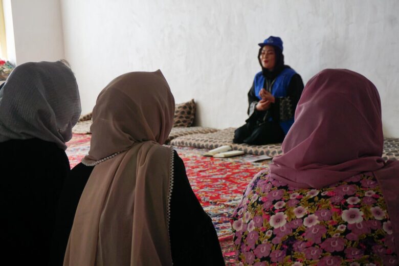 Three people in headcoverings of varying colour sit with the back facing the camera and look towards someone at the front o the room with a calm expression on their face.