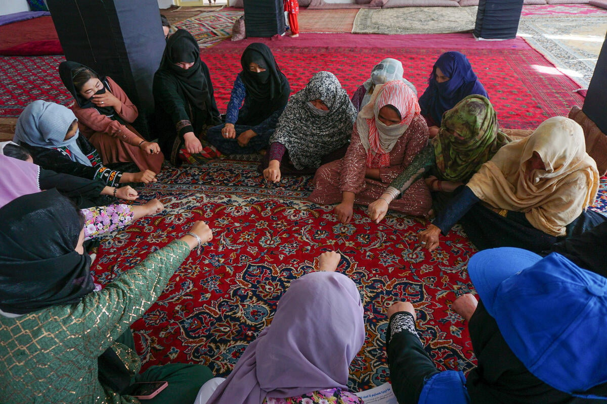 Several people sit on the floor in criss-cross position, each wearing headcoverings. They all reach into the center fo the circle they have gathered themselves in with their hands in a fist-like shape. 