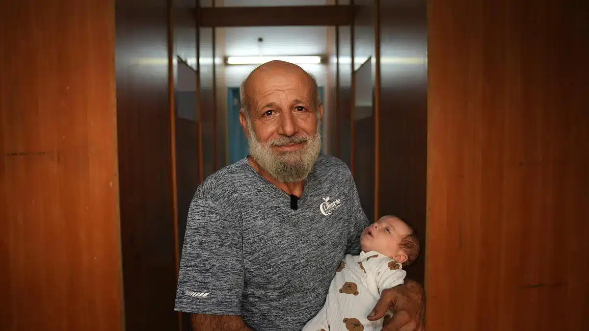 Person with gray facial hair gazes into the camera while holding an infant. The background is a hallway lined with wooden cupboards. The infant is wearing a white onesie with brown bear print displayed all over it and the adult is wearing a heather gray T-shirt. 