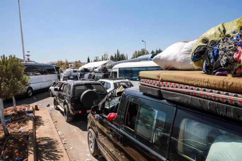 Several mid-sized vehicles and busses with belongings hastily strapped to the top, head the same direction on a road dusted with dirt and paper. There are trees in the horizon as well as on the bottom left corner, separating roadways. 